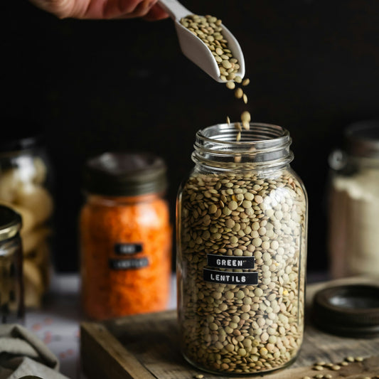 Person pouring green lentils into a jar labeled 'Green Lentils' on a wooden cutting board.