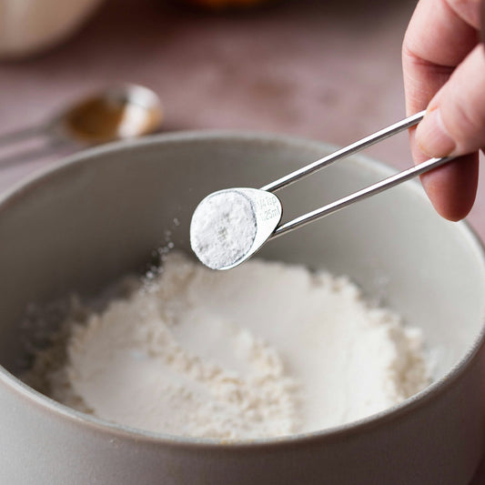 Hand using tongs to add salt to a bowl of flour with pumpkins in the background